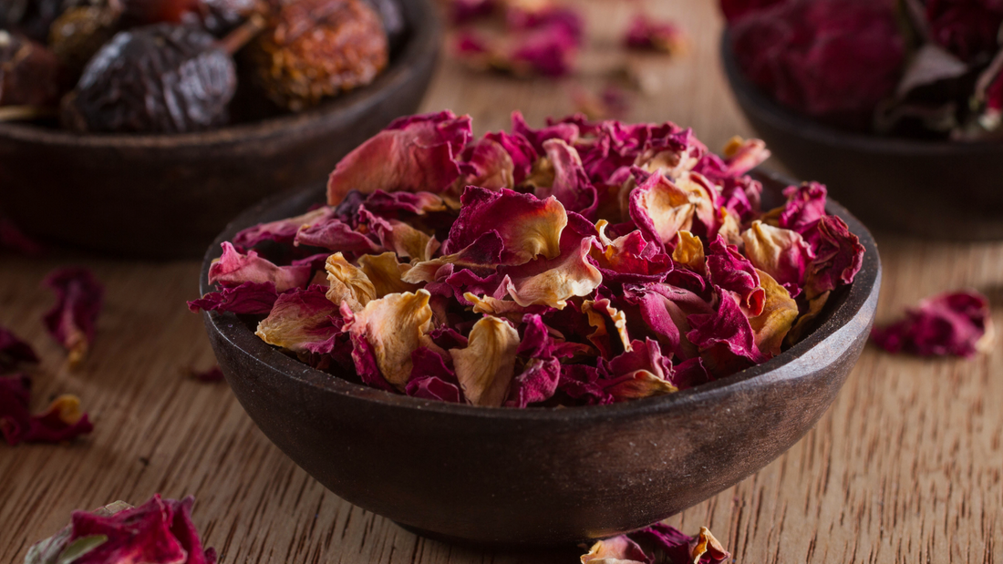 Close-up of dried rose petals in a dark wooden bowl on a rustic wood surface, used as a natural ingredient in skincare and apothecary formulations.