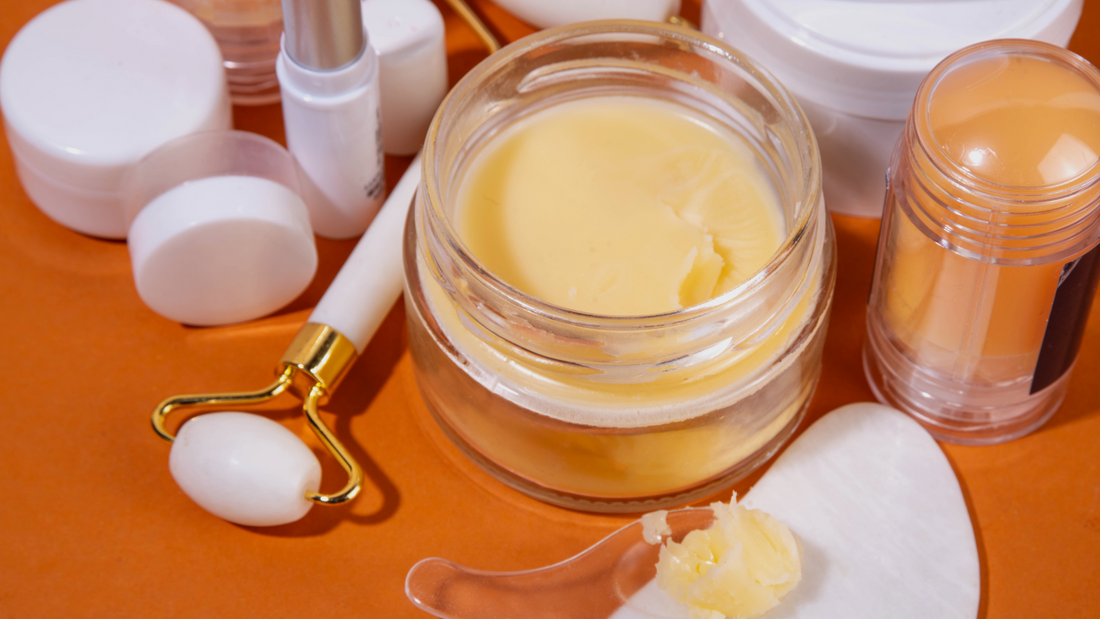 Glass jar of tallow balm surrounded by skincare tools and containers on an orange background, including a facial roller and spatula with balm, representing a natural skincare routine.