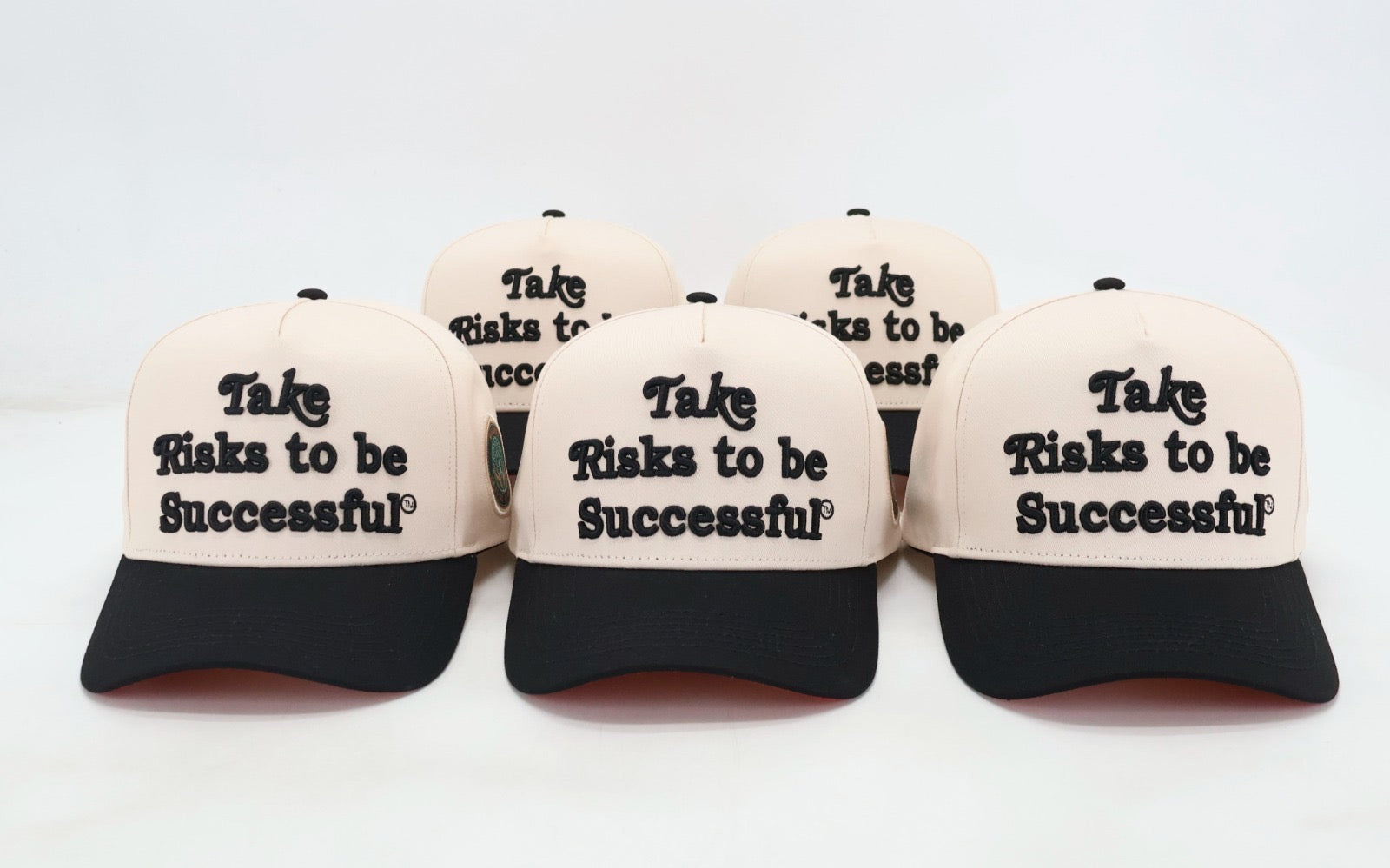A set of three baseball caps with the phrase 'Take Risks to Be Successful' printed on them, featuring a cream crown and a black visor.